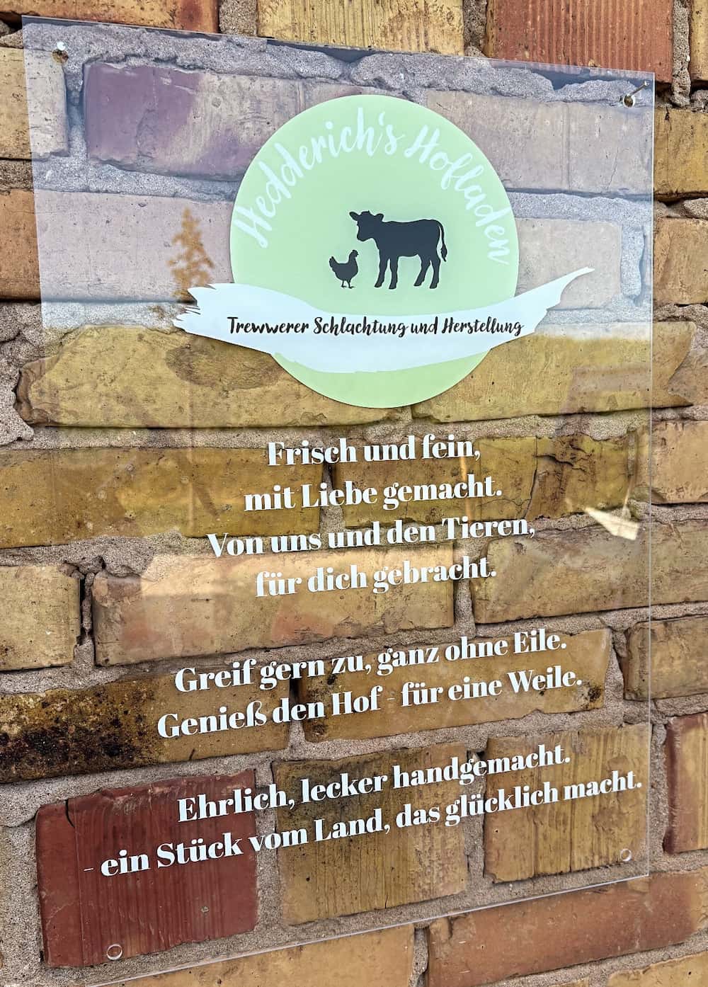 Ein Mann in formeller Kleidung hält stolz seinen Meisterbrief in der Hand. Im Hintergrund sind Kühe auf einer Wiese zu sehen, umgeben von einem Zaun. Der Standort deutet auf einen ländlichen Hofladen hin.